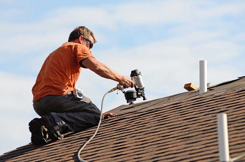 Homeowner examines a ceiling water stain while a roof with lifted shingles is visible through the attic hatch.