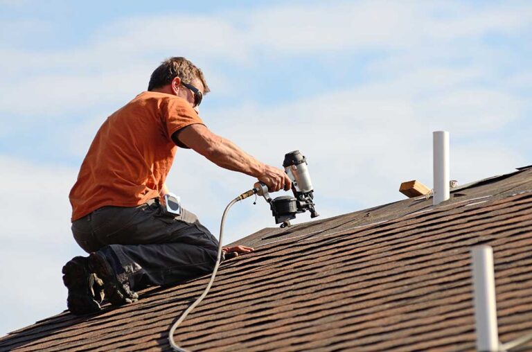 Homeowner examines a ceiling water stain while a roof with lifted shingles is visible through the attic hatch.
