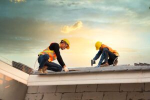 Roofing workers install shingles on a house while dry attic insulation is visible beneath the roof deck.
